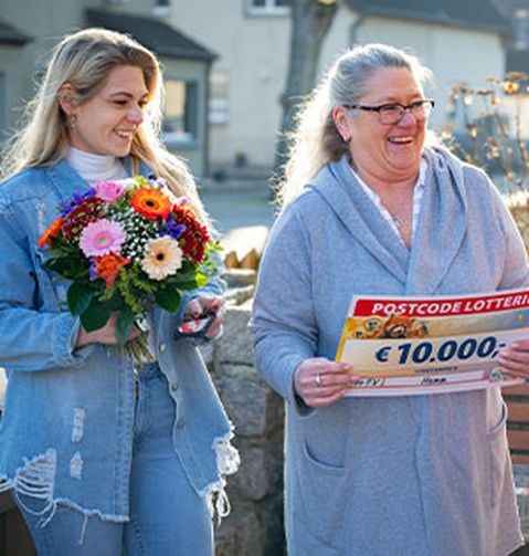 Zwei Frauen lächeln im Freien. Eine von ihnen hält einen Blumenstrauß, die andere einen großen Scheck über 10.000 € von der Postcode-Lotterie.
