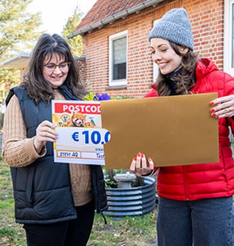 Zwei Frauen stehen vor einem Haus. Eine hält einen großen Umschlag, die andere ein Schild mit der Aufschrift „10.000 €“ und „Postcode“.